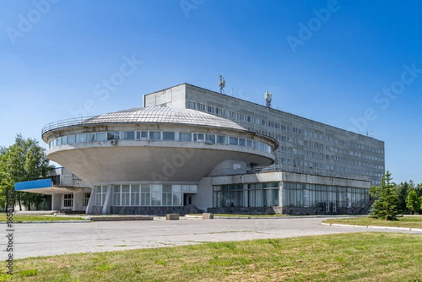 Fototapeta Technical university building in Ulyanovsk with a distinctive circular shape and concrete facade. Architectural structure in brutalist style under a clear blue sky surrounded by greenery.