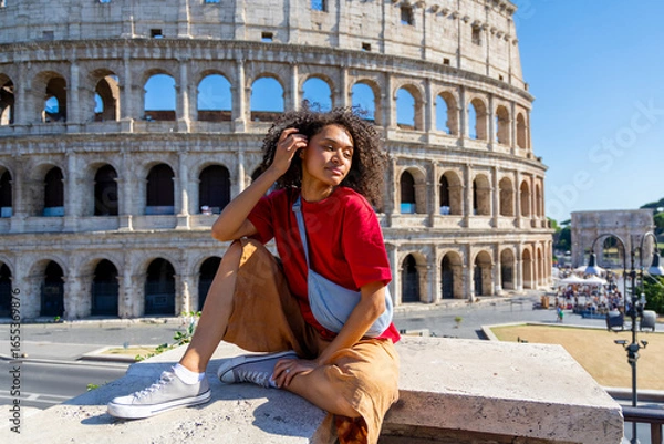 Fototapeta A stylish young woman elegantly sits on a ledge, gracefully posing while showcasing the iconic Colosseum in Rome, framed beautifully against a clear and vibrant blue sky that enhances the scene