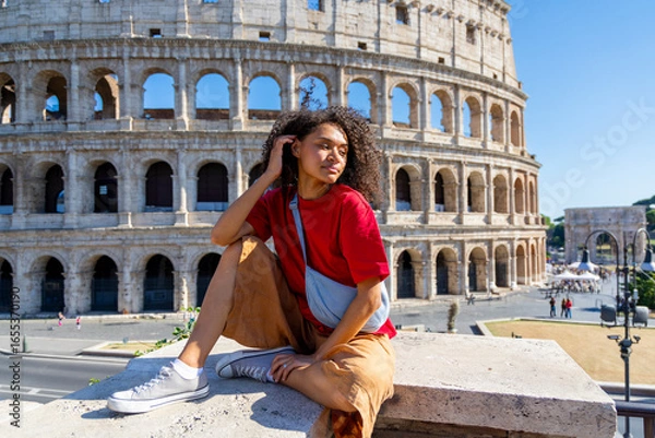 Fototapeta A stylish young woman in a chic outfit is sitting gracefully in front of the Colosseum, enjoying a sunny day while exploring Romes iconic historical site during her delightful adventure