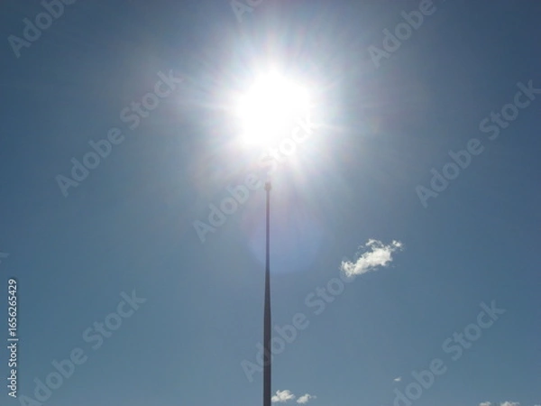 Obraz Bright sun flare above tall spire in blue sky
