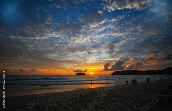 Fototapeta Golden Embrace above Kata beach.The setting sun paints the horizon with golden light, reflecting across gentle waves as silhouettes of beachgoers stroll peacefully along the shoreline.