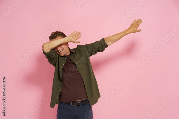 Fototapeta Joyful man performs the dab dance move in a studio with a plain pink background. Energetic expression captures playful mood of the moment, perfect for vibrant imagery.