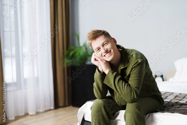 Fototapeta Happy young man sits comfortably on bed in pajamas, enjoying bright natural light streaming through the window. Scene conveys a sense of peace and relaxation during daytime.