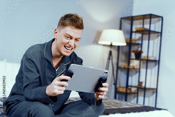Fototapeta Young man joyfully plays video game on tablet using controllers in cozy bedroom. Evening light creates a relaxed atmosphere filled with playfulness and excitement.