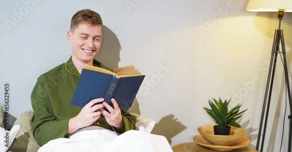 Fototapeta Happy young man immersed in reading a book in a cozy bedroom. Soft lighting from the floor lamp creates a peaceful atmosphere, enhancing the calm evening mood.