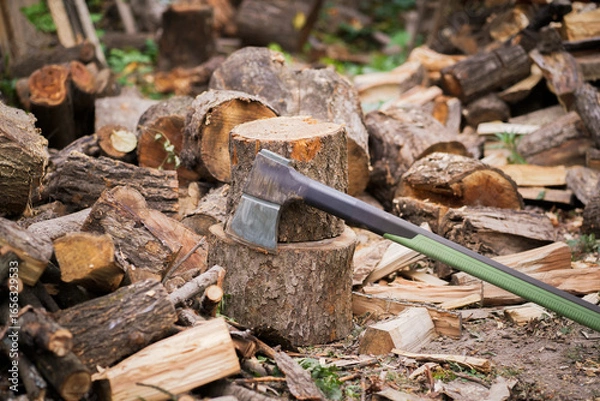 Fototapeta Rustic scene features an axe resting on a weathered tree stump. Surrounding pile of chopped firewood captures the essence of a utilitarian outdoor setting during the day.