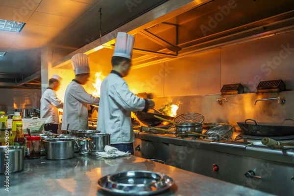 Fototapeta Szef kuchni w kuchni restauracji przy kuchence z patelnią, robi flambe na jedzeniu