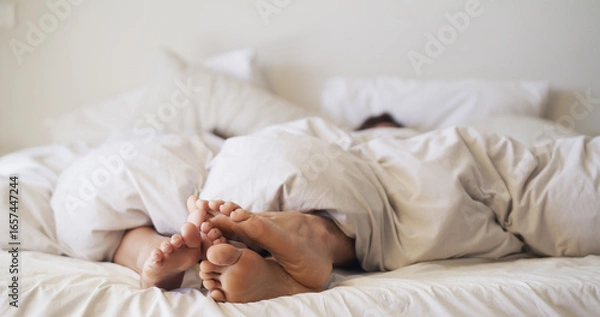 Fototapeta Feet, love and couple relax in bedroom for rest, peace and blanket for comfort in morning. Barefoot, people and touch in bed together on mattress with nap, sleeping and break on weekend in house