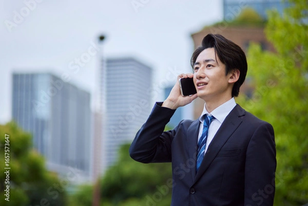 Fototapeta ビルを背景にスマートフォンで電話をするスーツを着た日本人ビジネスマンの男性