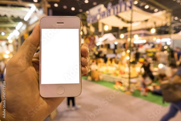 Fototapeta Holding mobile phone with de-focused shopping exhibition background, blank screen for text