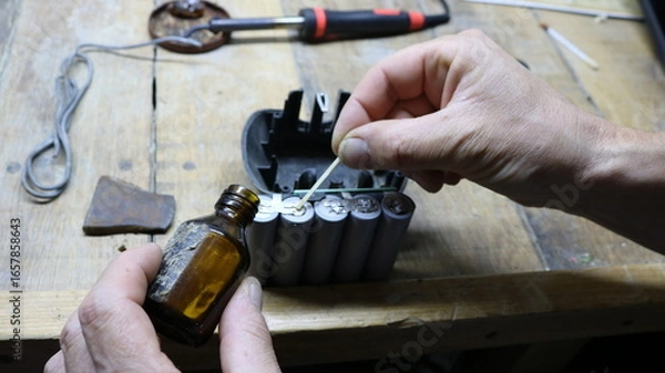 Fototapeta Processing contacts on old batteries from a construction tool during device resuscitation, close-up of male hands applying solution from a brown glass bottle to battery contacts on a work table
