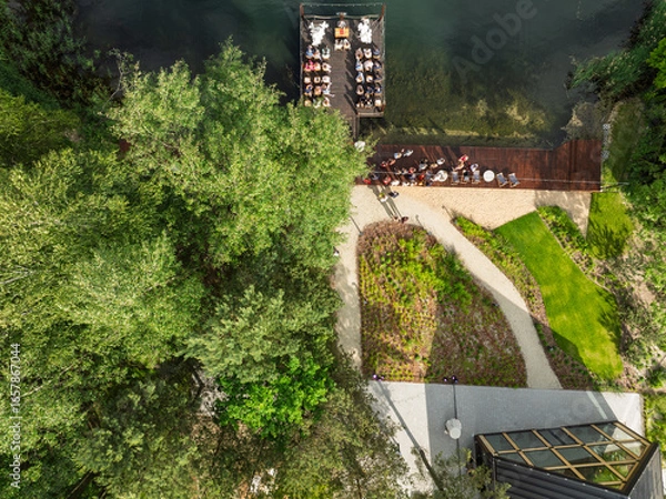 Fototapeta Aerial view of a wedding celebration by the lake, guests gathered on a wooden pier and terrace surrounded by lush green forest and a modern glass building in summer time.