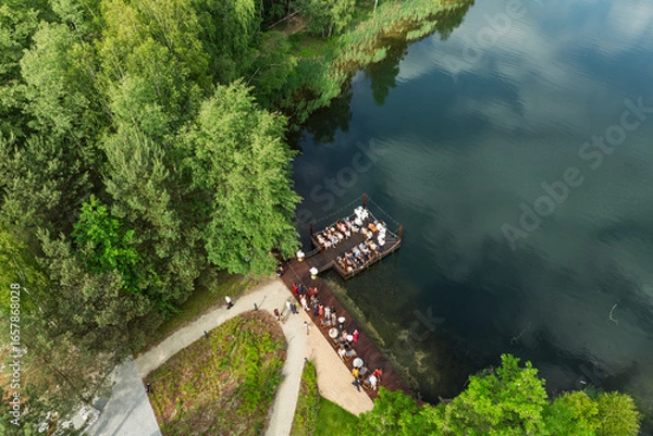 Fototapeta Aerial view of a wedding celebration by the lake, guests gathered on a wooden pier and terrace surrounded by lush green forest and a modern glass building in summer time.