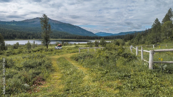 Fototapeta Tranquil mountain scene with a lake, lush greenery, and towering mountains. A dirt road, white vehicle, and dynamic clouds add to the serene atmosphere.