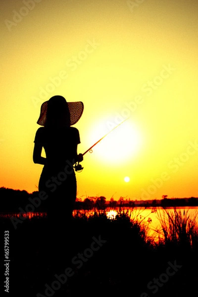 Fototapeta Silhouette of the figure of awoman in a hat with a fishing rod on the river bank
