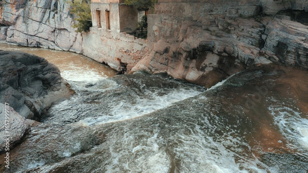 Fototapeta River with brown water and white foam flows through rocky channel, showcasing strong currents and turbulence. Dramatic scene from heightened perspective with trees in background.