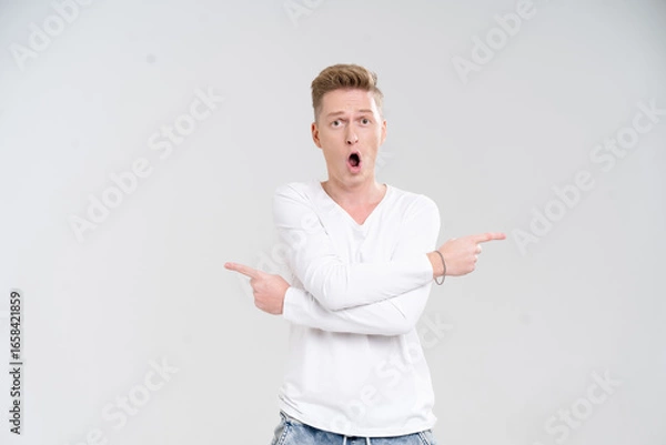 Fototapeta Young Caucasian man displays a surprised expression while pointing with both hands against a plain white background. His astonished demeanor captures a moment of excitement and wonder.