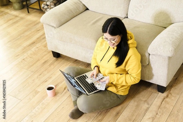 Fototapeta Happy Asian woman counts money while sitting in cozy living room during daytime. Joyful expression reflects satisfaction and contentment with finances.
