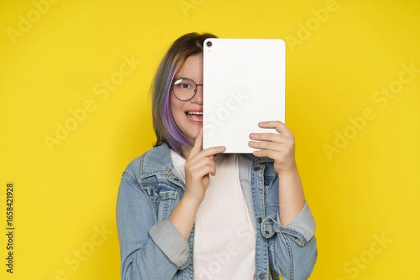 Fototapeta Happy mixed race girl holds tablet with excitement in bright studio. Yellow background enhances playful mood during daytime, showcasing her joyful expression.