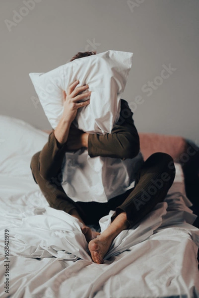 Fototapeta Young man, feeling anxious and stressed, hides face with a pillow in a dimly lit bedroom. Nighttime setting evokes feelings of loneliness and inner turmoil.