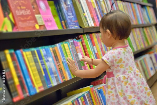 Obraz Azjatycka dziewczynka wybiera książkę w bibliotece. Dziecko patrzy na książki w bibliotece i decyduje, którą zabrać do domu. Kreatywność i wyobraźnia dzieci.