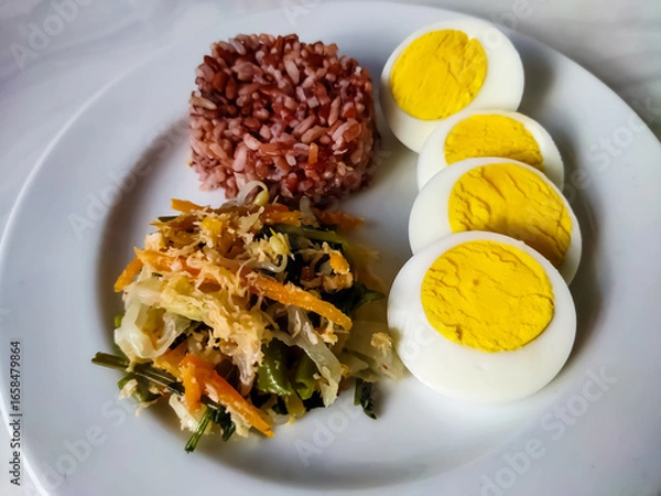 Fototapeta Red rice with vegetable salad or gudangan and a boiled egg on a white plate. A healthy meal concept.