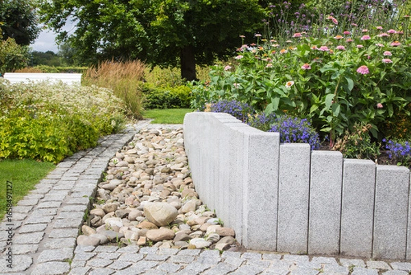 Fototapeta Modern Garden Design and Landscaping:Raised bed fenced by concrete blocks in different heights vertically erected-planted with flowers, perennial herbs-surrounded by grass, cobble and pebble stones