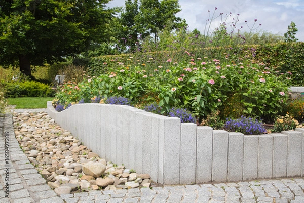 Fototapeta Modern Garden Design and Landscaping:Raised bed fenced by concrete blocks in different heights vertically erected-planted with flowers, perennial herbs-surrounded by grass, cobble and pebble stones