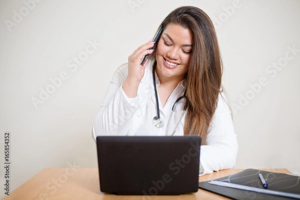 Fototapeta Happy smiling young female hispanic doctor sitting behind the desk and talking on the mobile phone