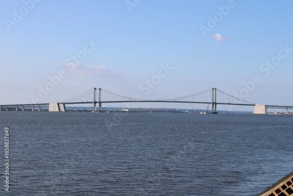 Obraz View of a Modern Suspension Bridge Spanning Across a Calm Body of Water Under a Clear Sky, Showcasing Engineering Marvel Against a Peaceful Landscape