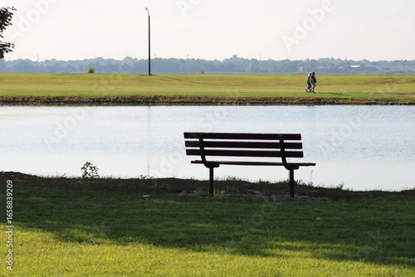 Obraz Peaceful Bench by the Lake With a Scenic View, Inviting Relaxation and Contemplation in the Afternoon Sun, Surrounded by Lush Greenery and Distant Walking Figures