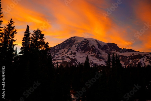 Fototapeta Stunning sunset over Mount Rainier, Washington. Vibrant sky with warm hues illuminating the rugged mountain peaks and surrounding forest.
