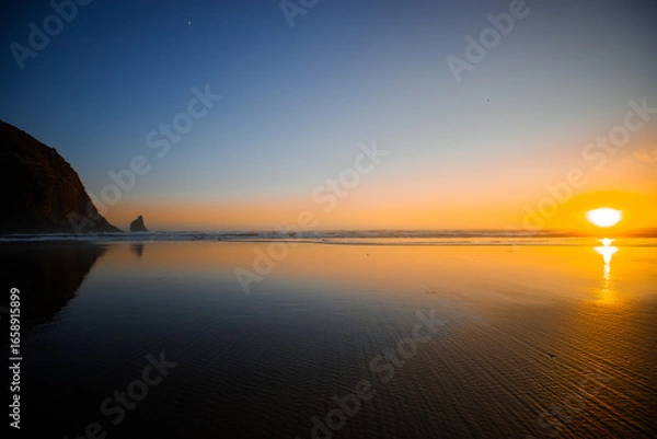 Fototapeta Sunset over Cannon Beach, Oregon. Vibrant sky, ocean waves, and the iconic Haystack Rock silhouetted against the evening light.