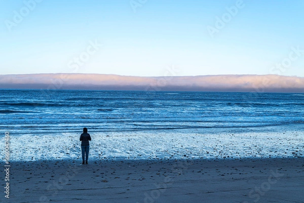 Fototapeta A person stands on an empty beach looking at the Pacific Ocean.