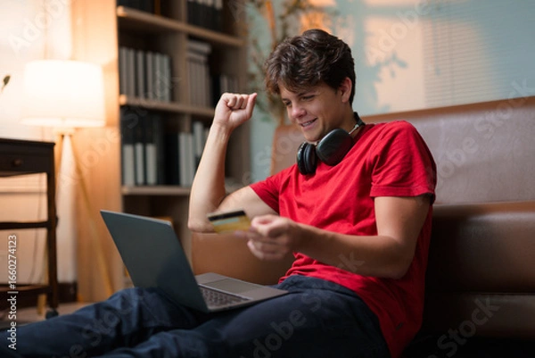 Fototapeta Happy teenager making online payment using credit card and laptop, celebrating successful purchase from home