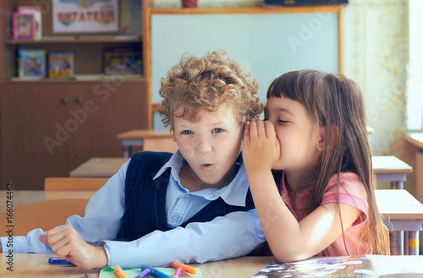 Fototapeta Girl telling secret to surprised boy in classroom.