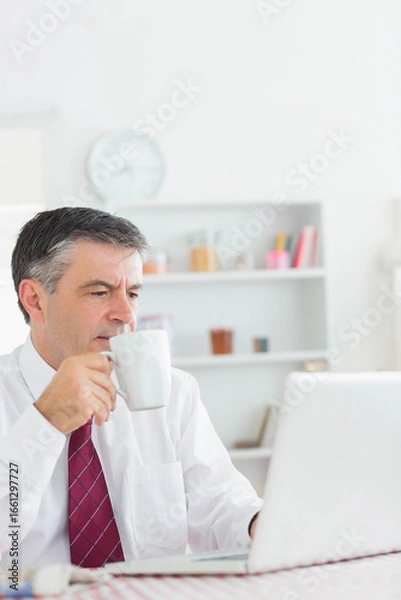 Fototapeta Middle-aged man holding white coffee mug, checking laptop at desk in home office, copy space