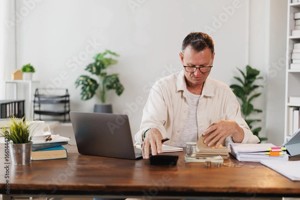Fototapeta Focused mature accountant managing budget, calculating home finances and making notes while working with laptop and financial documents at home office