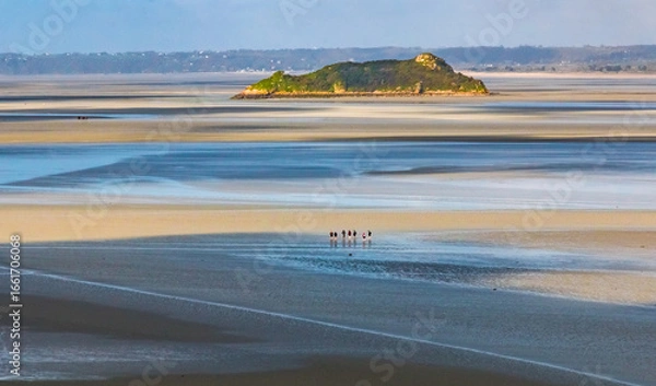 Fototapeta A picturesque scene stretches across the bay as a distant group of mudflat hikers wade over shimmering low‑tide sands toward the small tidal island of Tombelaine.