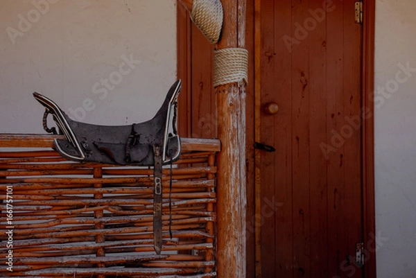 Fototapeta A rustic wooden house in the Caucasus region. The house features a woven fence and a saddle hanging on the porch. The window is partially open, revealing a glimpse of the interior.