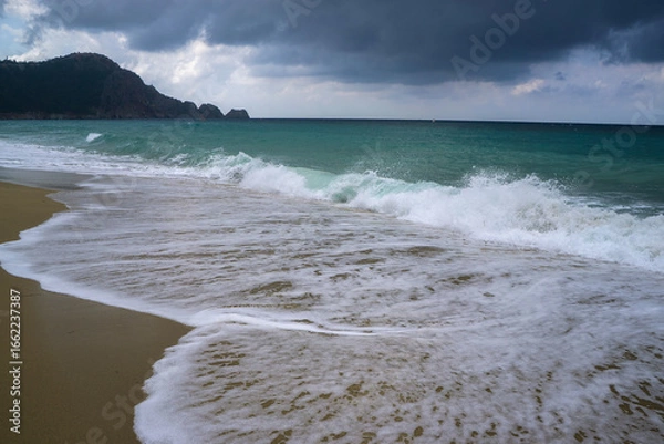 Fototapeta Waves crashing on sandy beach under cloudy sky