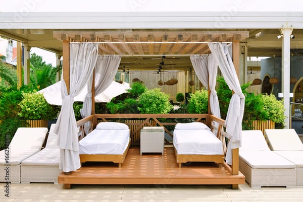 Obraz Cozy beach cabana with white beds and curtains surrounded by green plants at summer sunny day. Place for the rest on resort.