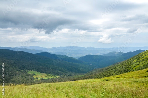 Fototapeta View of a mountain valley under a dramatic, cloudy sky. The foreground is a grassy meadow, while the distant mountains are layered in shades of blue and green. Carpathians, Ukraine