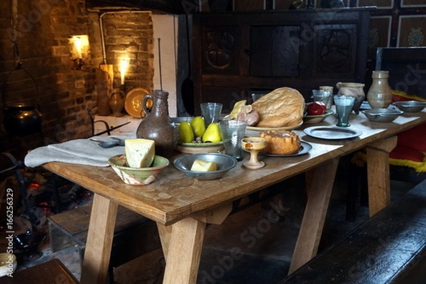 Obraz Rustic old wooden dining table in a medieval cottage, laid with bread, meat, cheese and fruit, and antique pottery and pewter