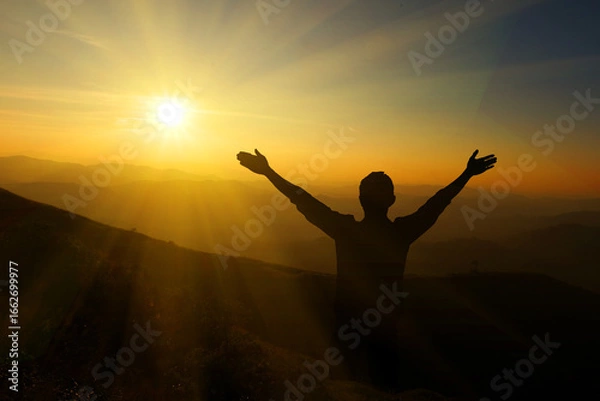 Fototapeta Silhouette of a man with raised hands on the top of the mountain.