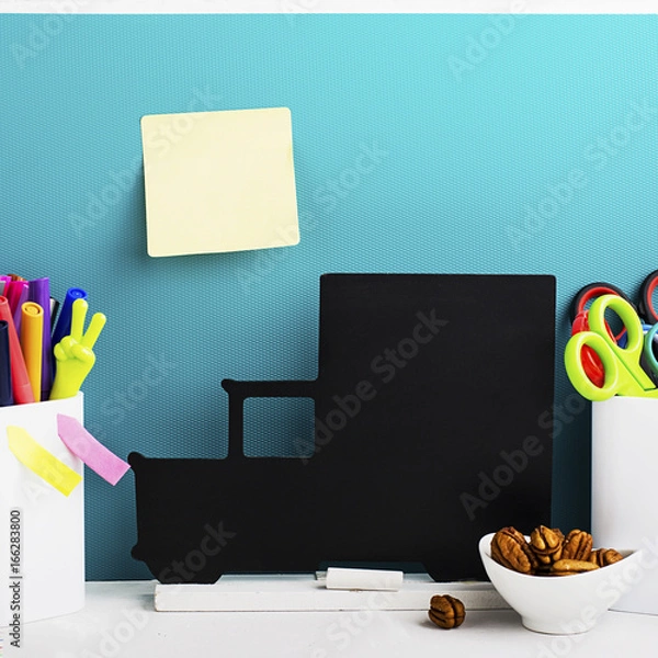Fototapeta School background: a white brick wall with accessories, pencils, tetrads, scissors, an apple, pecans, a board for a substitute placed in front of her on the table. Selective focus.