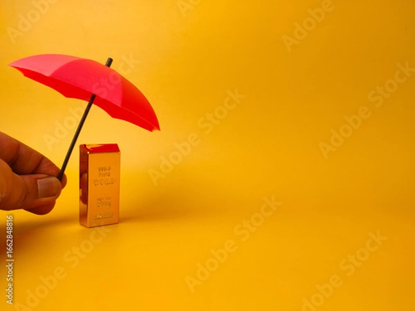 Fototapeta Someone hand holding red umbrella covered gold bar on a yellow background with copy space