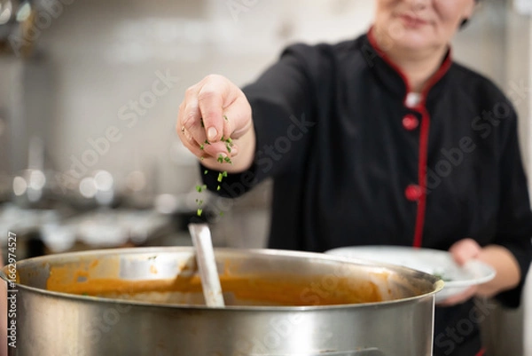 Fototapeta Close up of a chef in black uniform adding fresh herbs to a large pot of soup in a professional kitchen. Concept of gastronomy, seasoning, culinary preparation, and hospitality industry