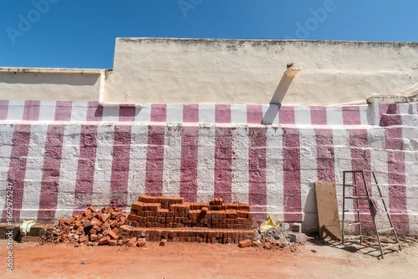 Fototapeta Brightly colored striped wall features stacked bricks in front, showcasing a construction area on a sunny day.