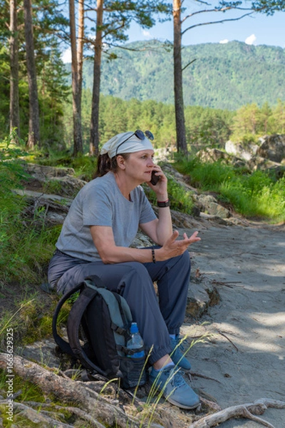 Obraz An adult attractive woman is sitting on the rocks on the bank of a mountain river and talking on the phone.
